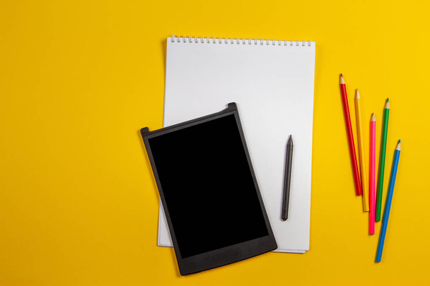 Tableta gráfica digital, lápices de colores, cuaderno de papel blanco sobre fondo amarillo
 - Foto, imagen