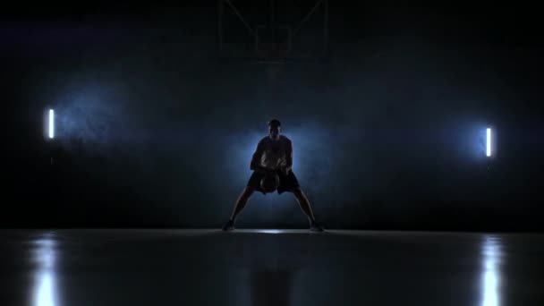 Een man met een basketbal op een donkere basketbalveld tegen de achtergrond van een basketbal-ring in de rook toont dribbelen vaardigheden verlicht door drie lantaarns in tegenlicht - Video