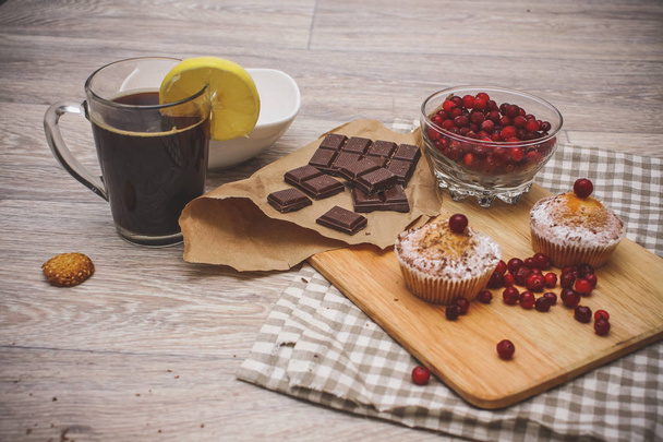 On a light wooden tabletop on a linen napkin napkin, there is a cutting board with two muffins, a broken chocolate bar and bright red berries in a small tree, next to a bowl with cookies. Festive pastry composition. - 写真・画像