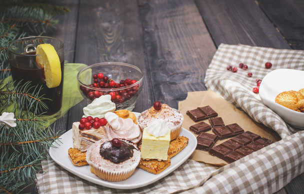 Close-up of sweets on a white plate: coconut biscuit, pastila, meringue, cream roses, Turkish delight, next to a broken chocolate bar on craft paper, in a bowl sesame biscuits in a bowl on a checkered linen napkin, fresh red cranberries, a glass of d - 写真・画像