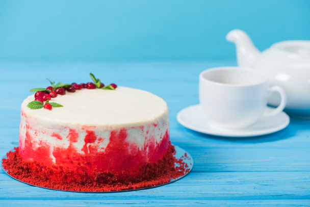 cake decorated with red currants and mint leaves near white cup and tea pot isolated on blue - Foto, Imagem