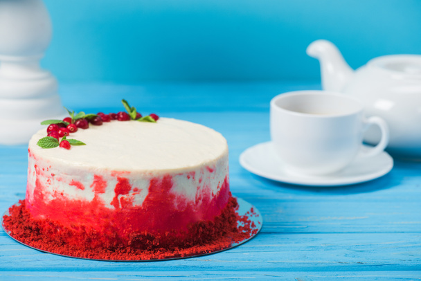 cake decorated with red currants and mint leaves between white cup, tea pot and stand isolated on blue - Photo, Image