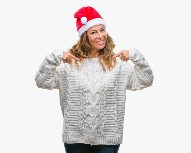 Mujer hispana mayor de mediana edad con sombrero de navidad sobre un fondo aislado que luce confiado con sonrisa en la cara, señalándose con los dedos orgullosos y felices
. - Foto, Imagen