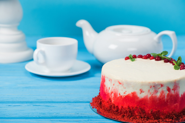 cake decorated with red currants and mint leaves between white cup, tea pot and stand isolated on blue - Фото, изображение