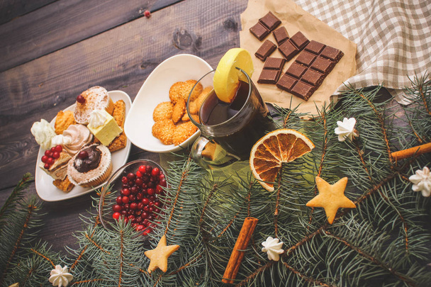 Happy New Year fir branches decorated with gingerbread stars, cinnamon sticks, dried orange slices and meringue peaks, bright cranberries, sweets in a dessert plate, a broken chocolate bar on kraft paper and a glass of tea with lemon on a linen napki - Valokuva, kuva