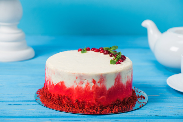 cake versierd met rode aalbessen en muntblaadjes tussen witte cup, theepot en stand geïsoleerd op blauw - Foto, afbeelding