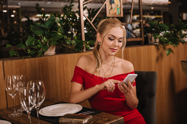 mulher atraente em vestido vermelho sentado no restaurante e usando smartphone
 - Foto, Imagem