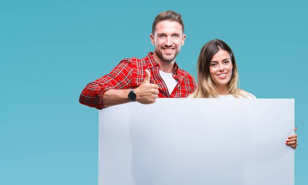 Young couple together holding blank banner over isolated background happy with big smile doing ok sign, thumb up with fingers, excellent sign - Photo, Image