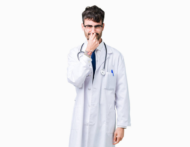 Νέος γιατρός άνδρας φοράει παλτό νοσοκομείο πέρα από απομονωμένες φόντο μυρίζουν κάτι stinky και αηδιαστικό, αφόρητη μυρωδιά, εκμετάλλευση αναπνοή με δάκτυλα στη μύτη. Άσχημες μυρωδιές έννοια. - Φωτογραφία, εικόνα