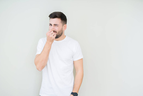 Jonge knappe man dragen casual wit t-shirt over geïsoleerde achtergrond iets ruiken stinkende en walgelijk, ondraaglijke geur, bedrijf adem met vingers op de neus. Slechte geuren concept. - Foto, afbeelding