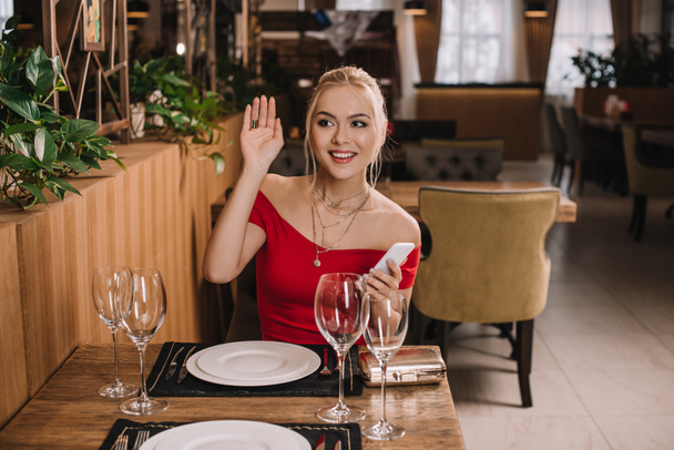 atractiva mujer en vestido rojo sentado en restaurante saludando la mano
 - Foto, Imagen