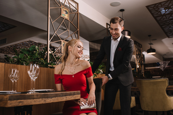 handsome man helping to sit down attractive woman in red dress and holding chair - Foto, imagen
