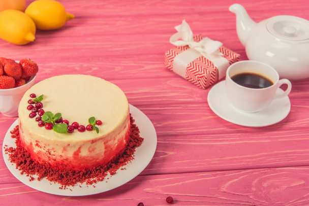 close up of cake decorated with currants and mint leaves near tea pot, cup of strawberries and gift box on pink surface - Foto, Imagen