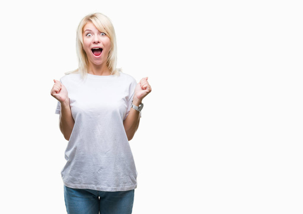 Young beautiful blonde woman wearing white t-shirt over isolated background celebrating surprised and amazed for success with arms raised and open eyes. Winner concept. - Foto, afbeelding