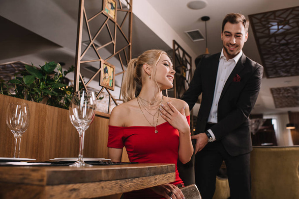 guapo novio ayudando a sentarse atractiva novia en vestido rojo, sonriendo y sosteniendo silla
 - Foto, Imagen