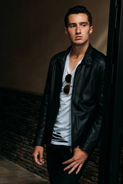 young, handsome guy in a white T-shirt, black, leather jacket and black pants is on the street, n the T-shirt sunglasses. model, city walk, live style - Foto, Imagen