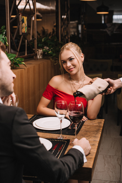 ragazza allegra sorridente al fidanzato mentre il cameriere versa il vino rosso nel bicchiere nel ristorante
  - Foto, immagini