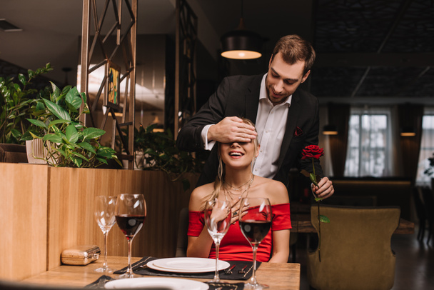 ωραίος ο φίλος, κλείνοντας τα μάτια του ελκυστική κοπέλα σε κόκκινο φόρεμα, κρατώντας τριαντάφυλλο  - Φωτογραφία, εικόνα