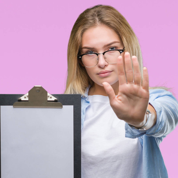 Young caucasian business woman holding clipboard over isolated background with open hand doing stop sign with serious and confident expression, defense gesture - Foto, imagen