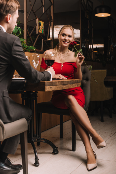 ελκυστική κοπέλα κάθεται με κόκκινο τριαντάφυλλο και κοιτάζοντας φίλο στο εστιατόριο  - Φωτογραφία, εικόνα