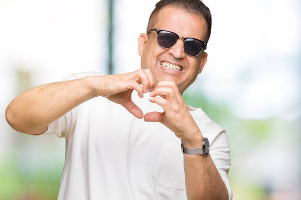 Średnim wieku Emiraty człowiek wearig biały t-shirt i okulary na białym tle uśmiechający się w miłość serce symbol i kształt rękami. Romantyczna koncepcja. - Zdjęcie, obraz