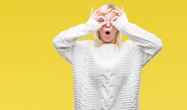 Young beautiful blonde woman wearing winter sweater and glasses over isolated background doing ok gesture like binoculars sticking tongue out, eyes looking through fingers. Crazy expression. - Foto, afbeelding