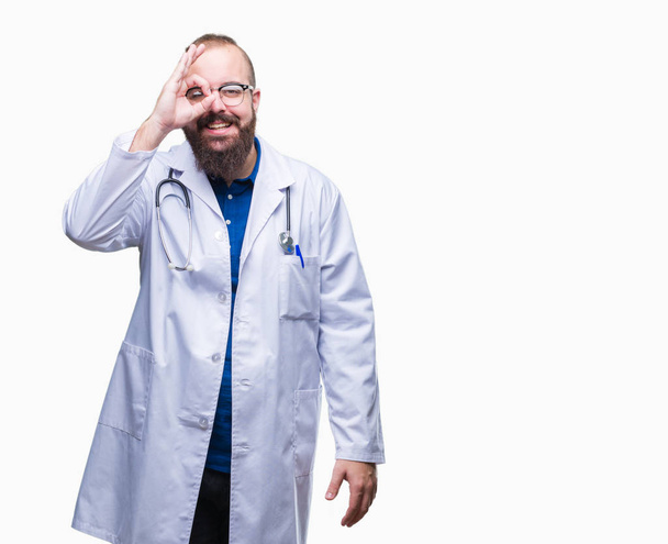 Young caucasian doctor man wearing medical white coat over isolated background doing ok gesture with hand smiling, eye looking through fingers with happy face. - Photo, Image