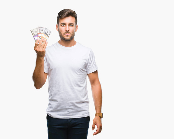 Young handsome man holding stack of dollars over isolated background with a confident expression on smart face thinking serious - Zdjęcie, obraz