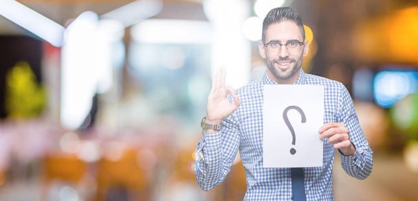 Handsome young business man holding paper with question mark over isolated background doing ok sign with fingers, excellent symbol - Photo, Image