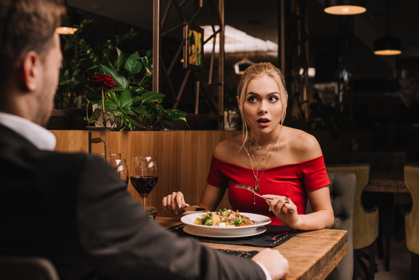 shocked woman looking at boyfriend while holding cutlery near salad in restaurant - Photo, Image