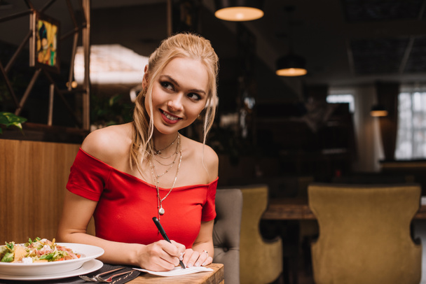 χαρούμενα ξανθιά γυναίκα γραψίματος σε χαρτί και να χαμογελά στο εστιατόριο  - Φωτογραφία, εικόνα