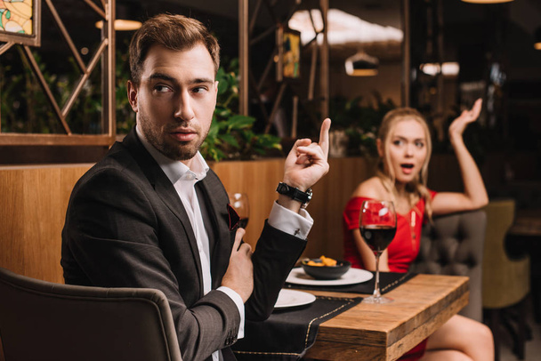 selective focus of offended man showing middle finger to shocked girlfriend in restaurant during dinner  - 写真・画像