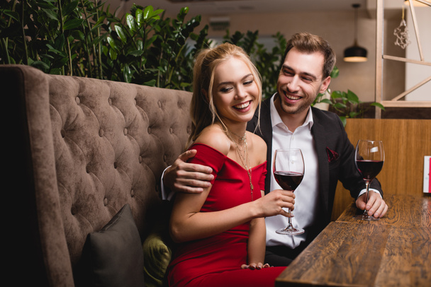 glückliches Mädchen lacht neben Freund, während es Glas mit Rotwein im Restaurant hält  - Foto, Bild