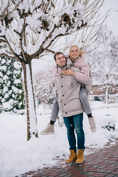cheerful man piggybacking smiling girlfriend on snowy street  - 写真・画像