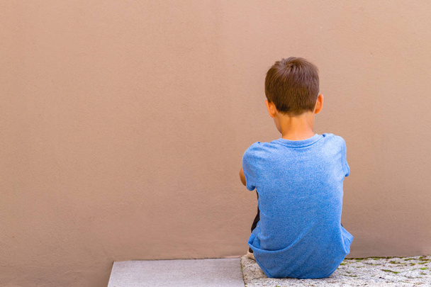 Triest alleen jongen zittend op de grond achter de muur buiten - Foto, afbeelding
