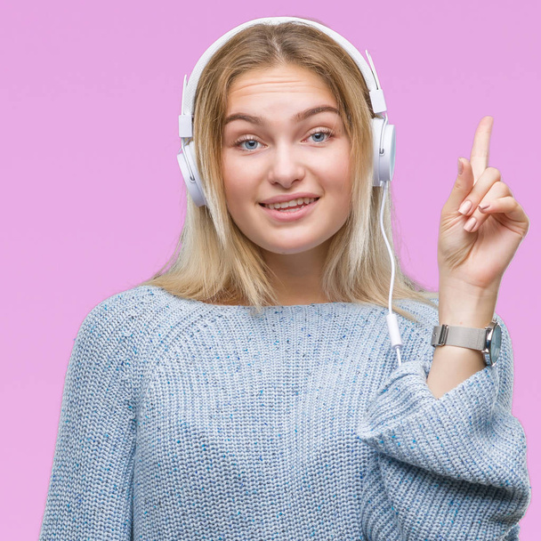 Jeune femme caucasienne écoutant de la musique portant des écouteurs sur fond isolé pointant du doigt avec une idée réussie. Sorti et heureux. Numéro un
. - Photo, image