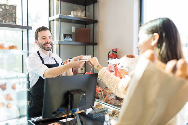 Happy salesman receiving credit card from customer in bakery - Фото, изображение