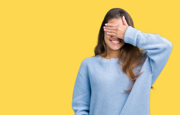 Young beautiful brunette woman wearing blue winter sweater over isolated background smiling and laughing with hand on face covering eyes for surprise. Blind concept. - Fotó, kép
