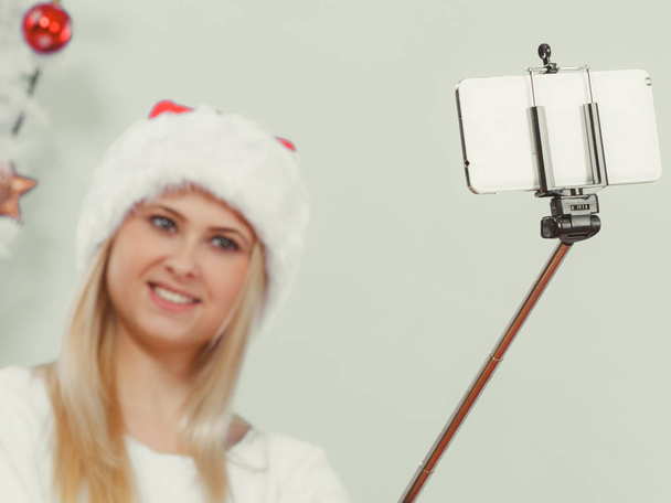 Modern holiday concept. Blonde woman in santa hat taking Christmas picture of herself, selfie with smartphone on stick. - Foto, Imagem