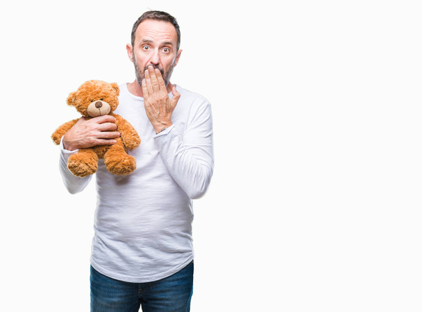 Middle age senior hoary man holding teddy bear over isolated background cover mouth with hand shocked with shame for mistake, expression of fear, scared in silence, secret concept - Valokuva, kuva