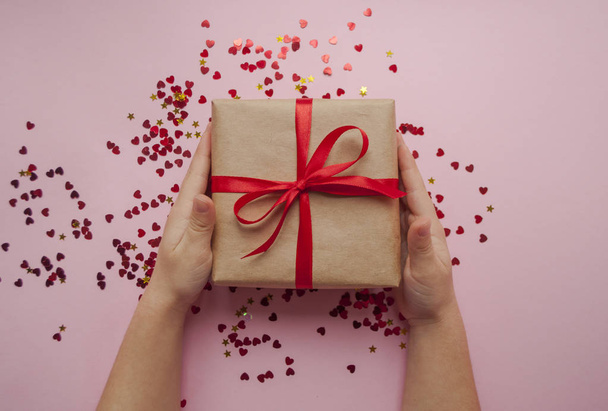 Child is holding gift box wrapped in brown colored craft paper and tied with red bow on pink background with red heart shape confetti. Flat lay style. - Φωτογραφία, εικόνα