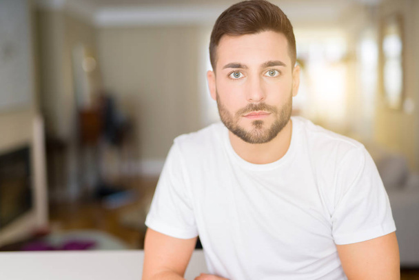 Young handsome man wearing casual white t-shirt at home with serious expression on face. Simple and natural looking at the camera. - Photo, Image