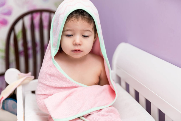 Niedliche weibliche Kleinkind trägt Kapuzenhandtuch, während sie im Kinderbett sitzt - Foto, Bild