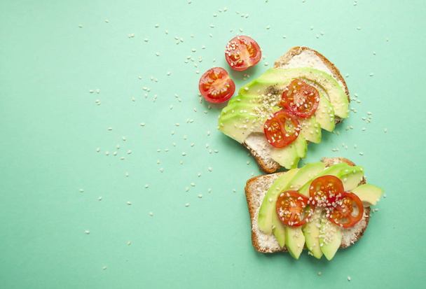 Flat lay of two delicious toasts with sliced avocado, tomatoes and sesamum seeds on turquoise background with copyspace. Healthy food concept. - Фото, изображение