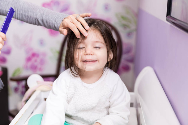 Jolie enfant souriant tandis que la mère toilettant ses cheveux à la maison
 - Photo, image