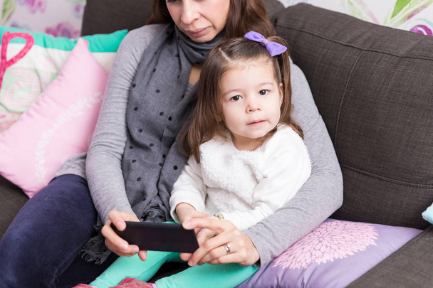 Portrait de fille mignonne assis avec la mère en utilisant le téléphone portable dans le salon à la maison
 - Photo, image