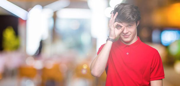 Mladý pohledný muž na sobě červené tričko izolované pozadí dělá ok gesto rukou, s úsměvem, oko dívá skrze prsty s šťastný obličej. - Fotografie, Obrázek