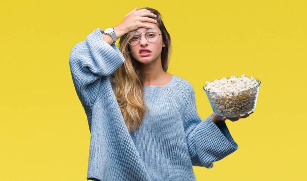Young beautiful blonde woman eating popcorn over isolated background stressed with hand on head, shocked with shame and surprise face, angry and frustrated. Fear and upset for mistake. - Fotoğraf, Görsel