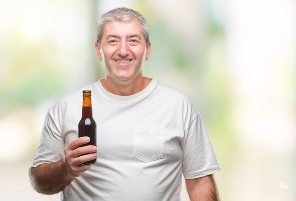 Bonito homem sênior bebendo garrafa de cerveja sobre fundo isolado com um rosto feliz de pé e sorrindo com um sorriso confiante mostrando dentes
 - Foto, Imagem