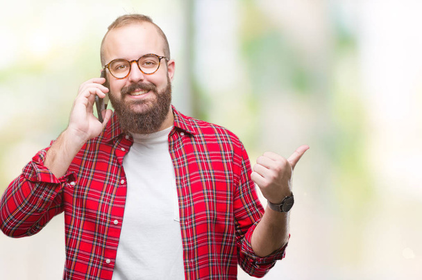 Jeune homme hipster caucasien parlant sur smartphone sur fond isolé pointant et montrant avec le pouce sur le côté avec sourire heureux visage
 - Photo, image